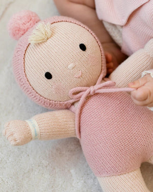A Cuddle + Kind My First Baby doll, hand-knit from natural cotton, lies on a soft surface beside a baby’s arm. The doll features a light hair tuft, a pink hood with pom-pom, and a hospital wristband on its arm.