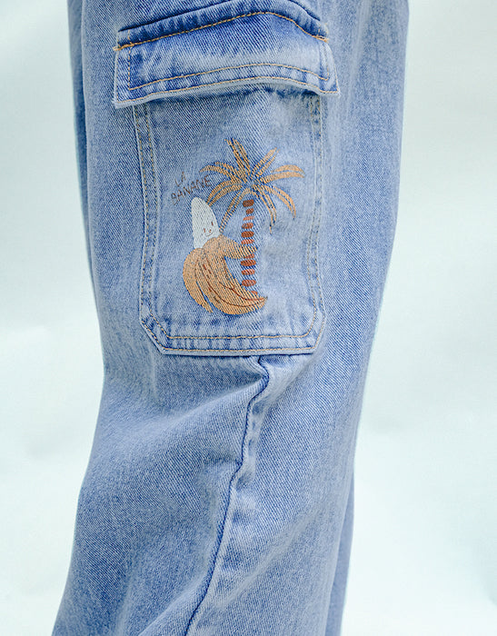 Gingersnaps Boys' Loose Cargo Pant Denim features a banana print and wide-leg cargo style in light blue, with an embroidered design on the pocket showing a palm tree, wave, and small white detail.