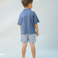 A young boy faces away, wearing a blue polo and Gingersnaps Boys' Pablo Twill Baltik Tile Drawstring Shorts with an elastic waist, paired with dark sandals against a light, neutral background.