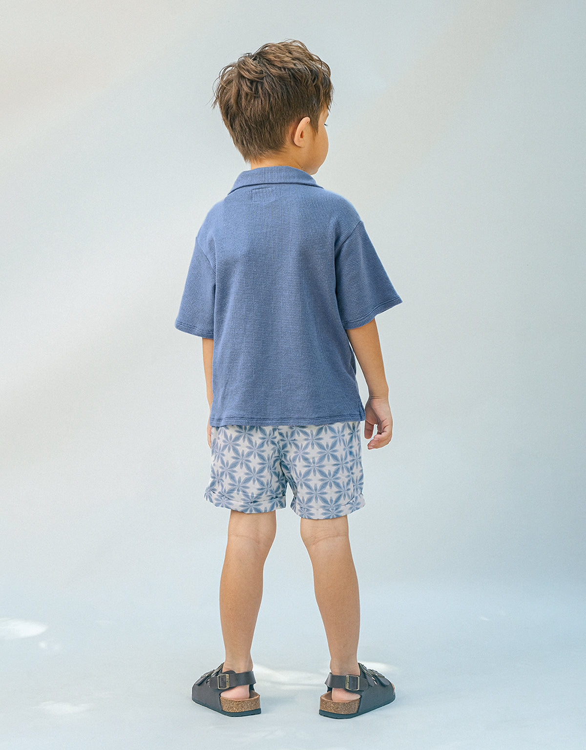 A young boy faces away, wearing a blue polo and Gingersnaps Boys' Pablo Twill Baltik Tile Drawstring Shorts with an elastic waist, paired with dark sandals against a light, neutral background.
