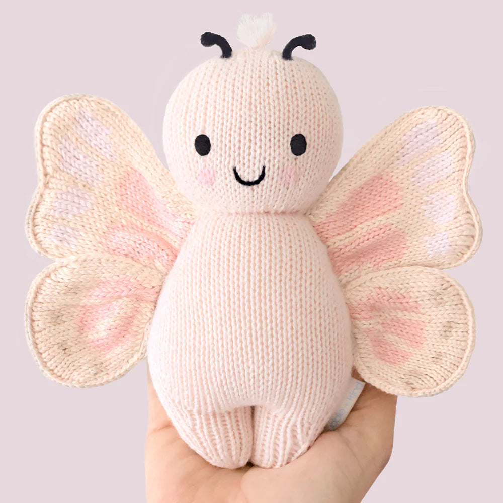 A hand holds the Cuddle + Kind Baby Butterfly - Petal Pink, an heirloom-quality plush toy with embroidered eyes, a smiling mouth, and patterned wings—a unique collectible for fans of hand-knit baby animals.