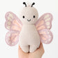 A hand holds the Baby Butterfly plush by Cuddle + Kind, crafted from Peruvian cotton yarn with pink and purple wings, black antennae, and a smiling face, shown against a neutral background.