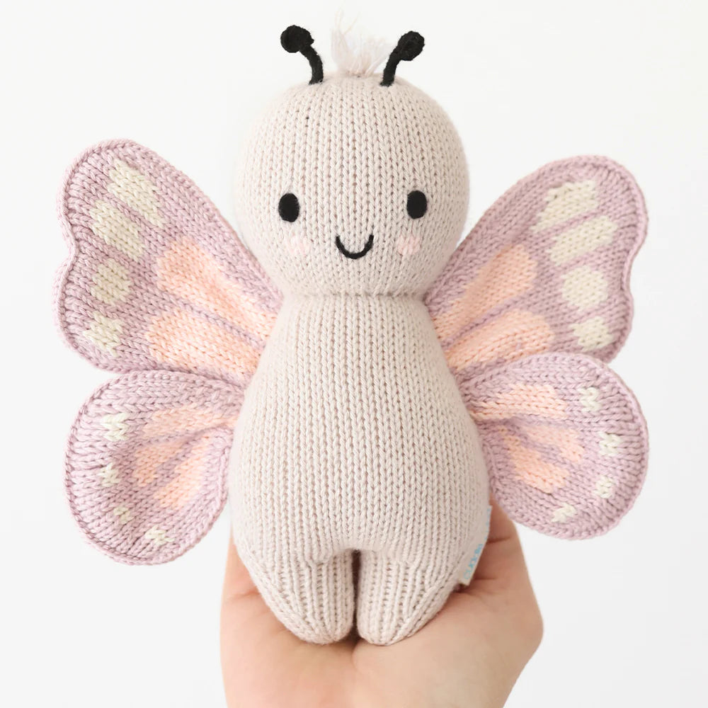 A hand holds the Baby Butterfly plush by Cuddle + Kind, crafted from Peruvian cotton yarn with pink and purple wings, black antennae, and a smiling face, shown against a neutral background.