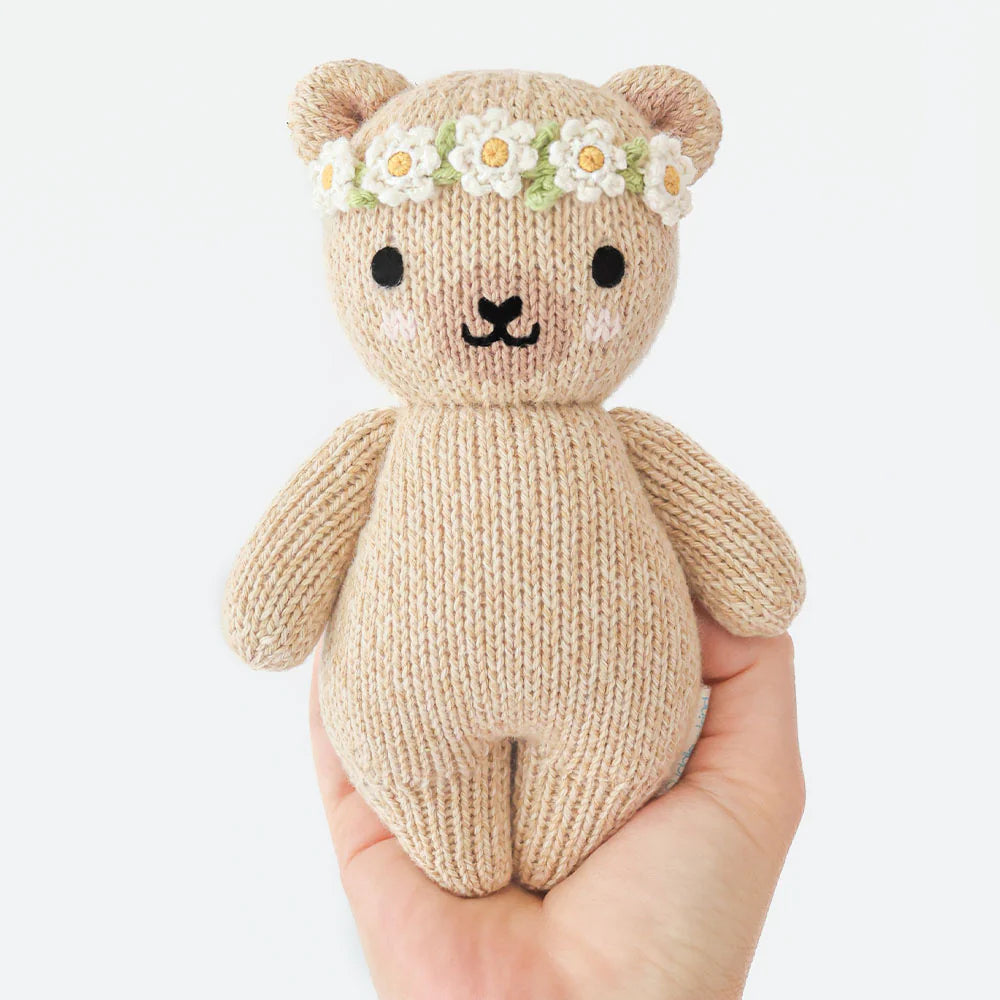 A hand holds the Baby Honey Bear Ivory Floral by Cuddle + Kind—an heirloom-quality, hand-knit beige teddy bear with a white and yellow flower crown and simple embroidered face.