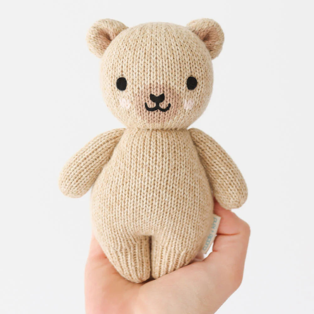 A hand holds the Baby Honey Bear by Cuddle + Kind—an heirloom-quality, beige hand-knit teddy crafted from Peruvian cotton yarn, featuring black eyes, a black nose, and blushing cheeks against a plain white background.