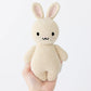 A hand holds Cuddle + Kind’s Baby Rabbit - Oatmeal, an heirloom-quality beige knitted bunny with black eyes, a simple smile, and upright ears against a plain white background.