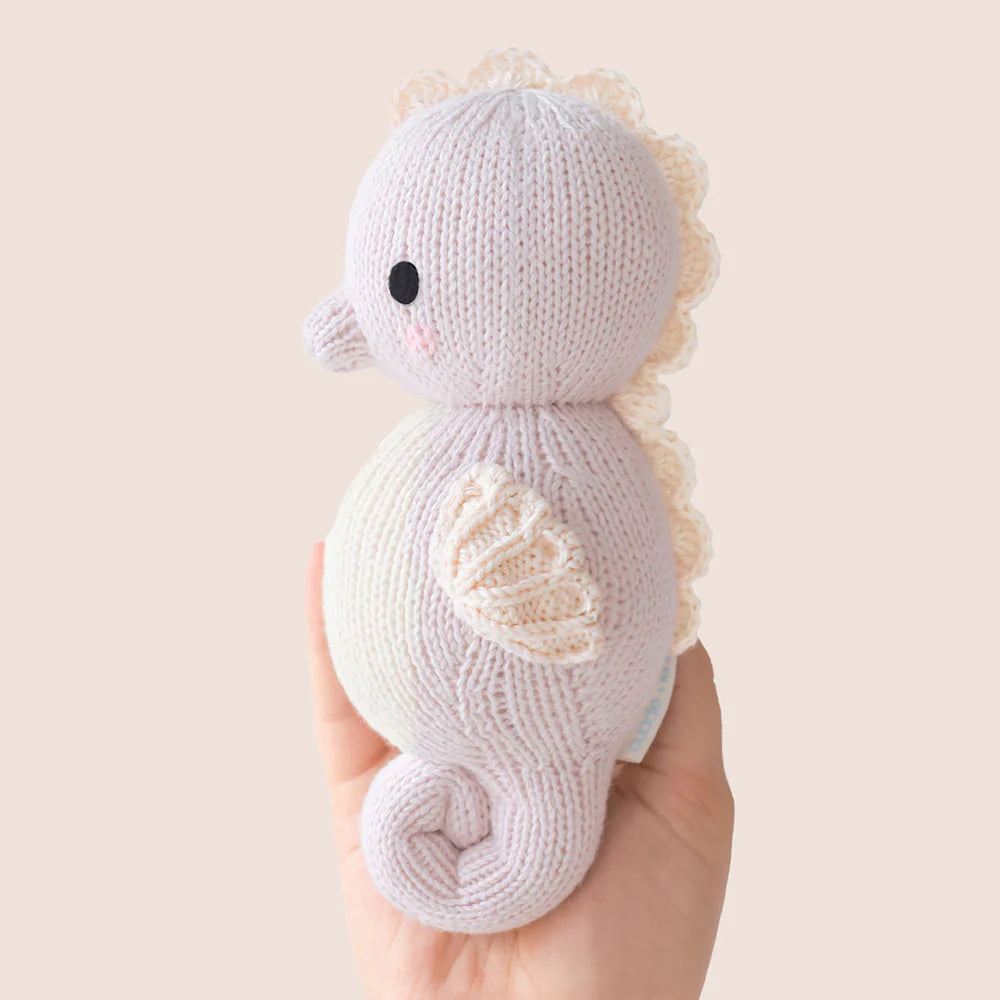 A hand holds the Baby Seahorse Lilac by Cuddle + Kind, an heirloom-quality, hand-knit toy in pastel lilac with a round body, small wings, decorative back edging, and crafted from soft Peruvian cotton yarn.