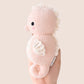 A hand holds the Cuddle + Kind Baby Seahorse - Powder Pink, an heirloom-quality, hand-knit toy with white accents and a curled tail. Made from soft Peruvian cotton yarn, it features black eyes against a plain light background.
