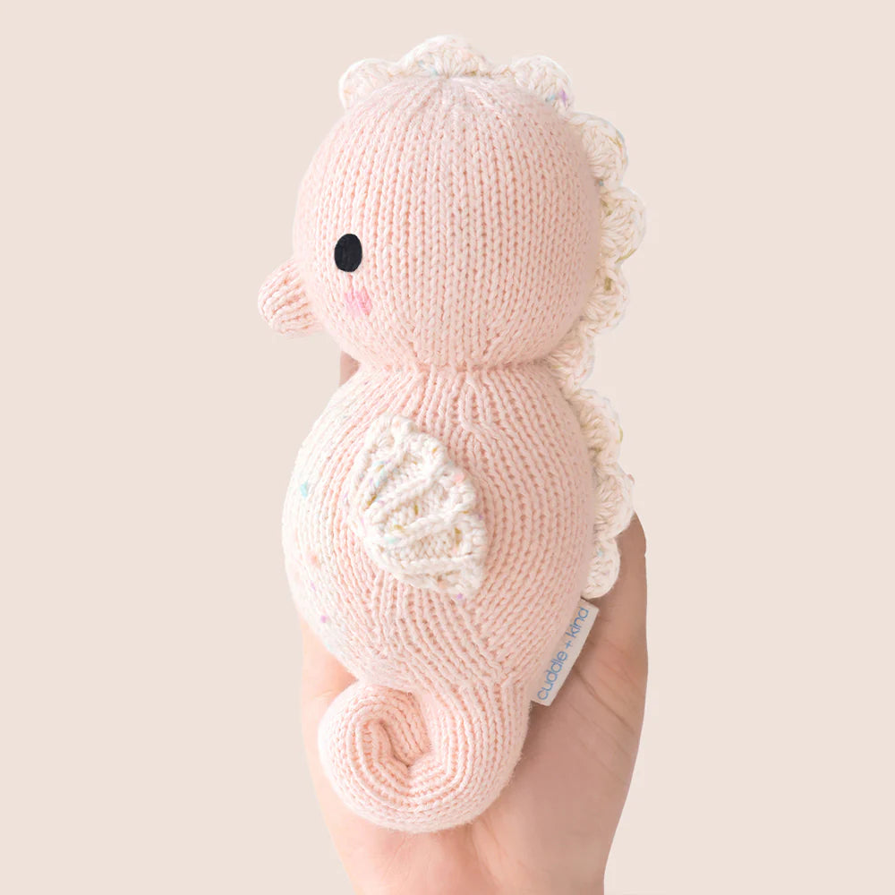 A hand holds the Cuddle + Kind Baby Seahorse - Powder Pink, an heirloom-quality, hand-knit toy with white accents and a curled tail. Made from soft Peruvian cotton yarn, it features black eyes against a plain light background.