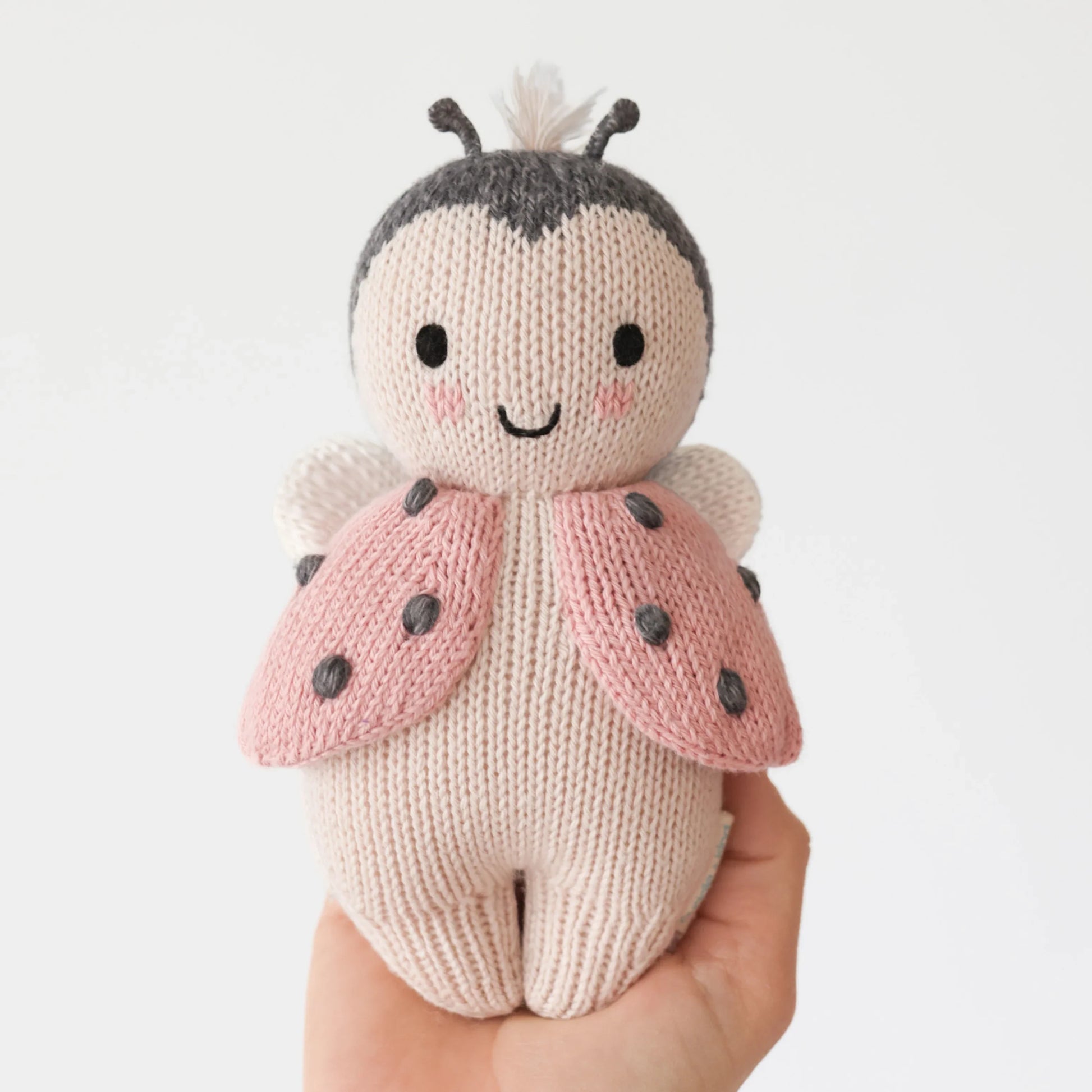 A hand holds Cuddle + Kind’s Baby Ladybug, an heirloom-quality, hand-knit plush toy with pink wings, black spots, and a smiling face, displayed against a plain white background.