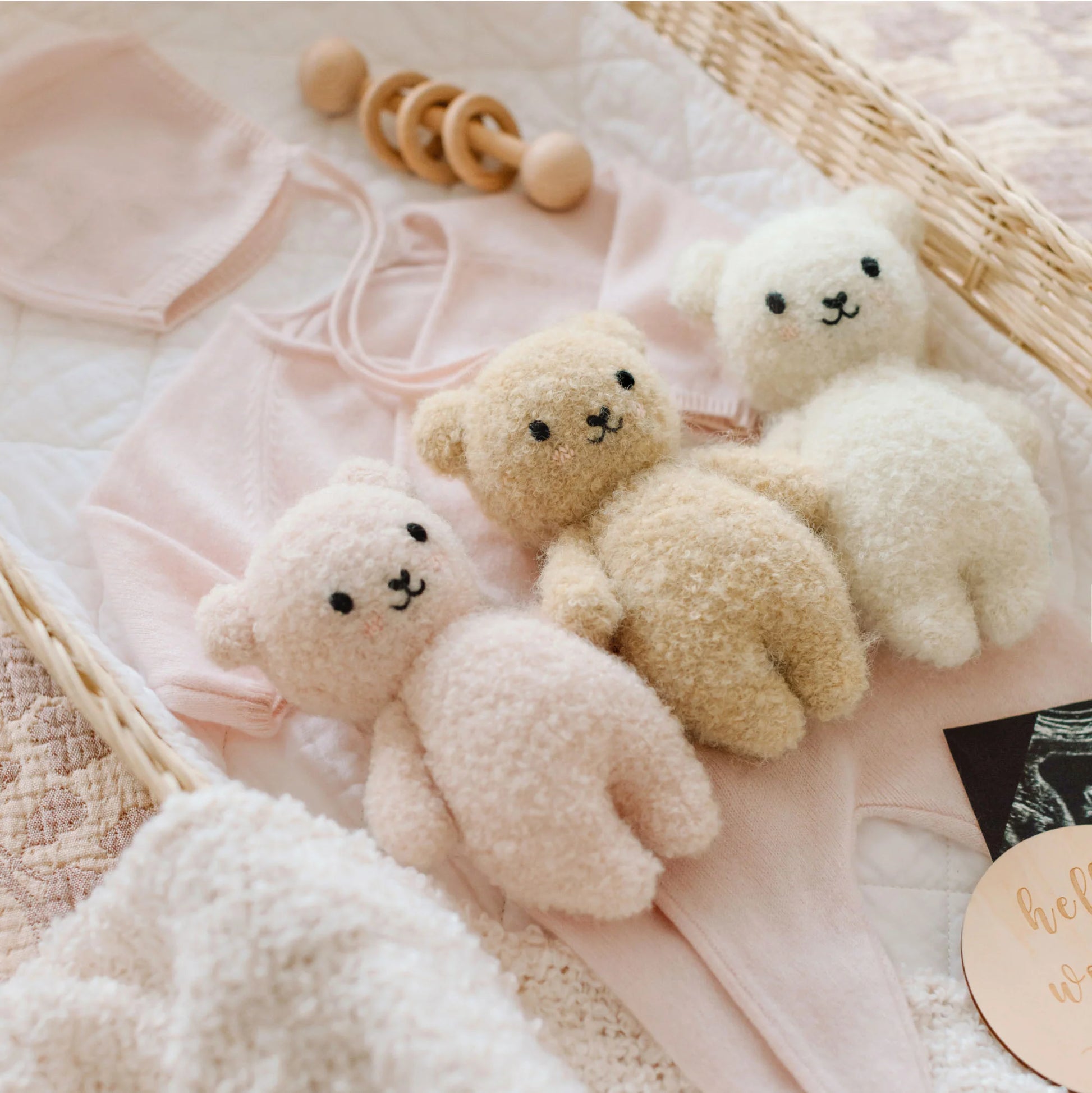 A Cuddle + Kind Baby Boucle Bear in Honey, crafted from Peruvian cotton yarn, is arranged on a pink baby outfit inside a wicker basket with a wooden rattle and other soft baby items.