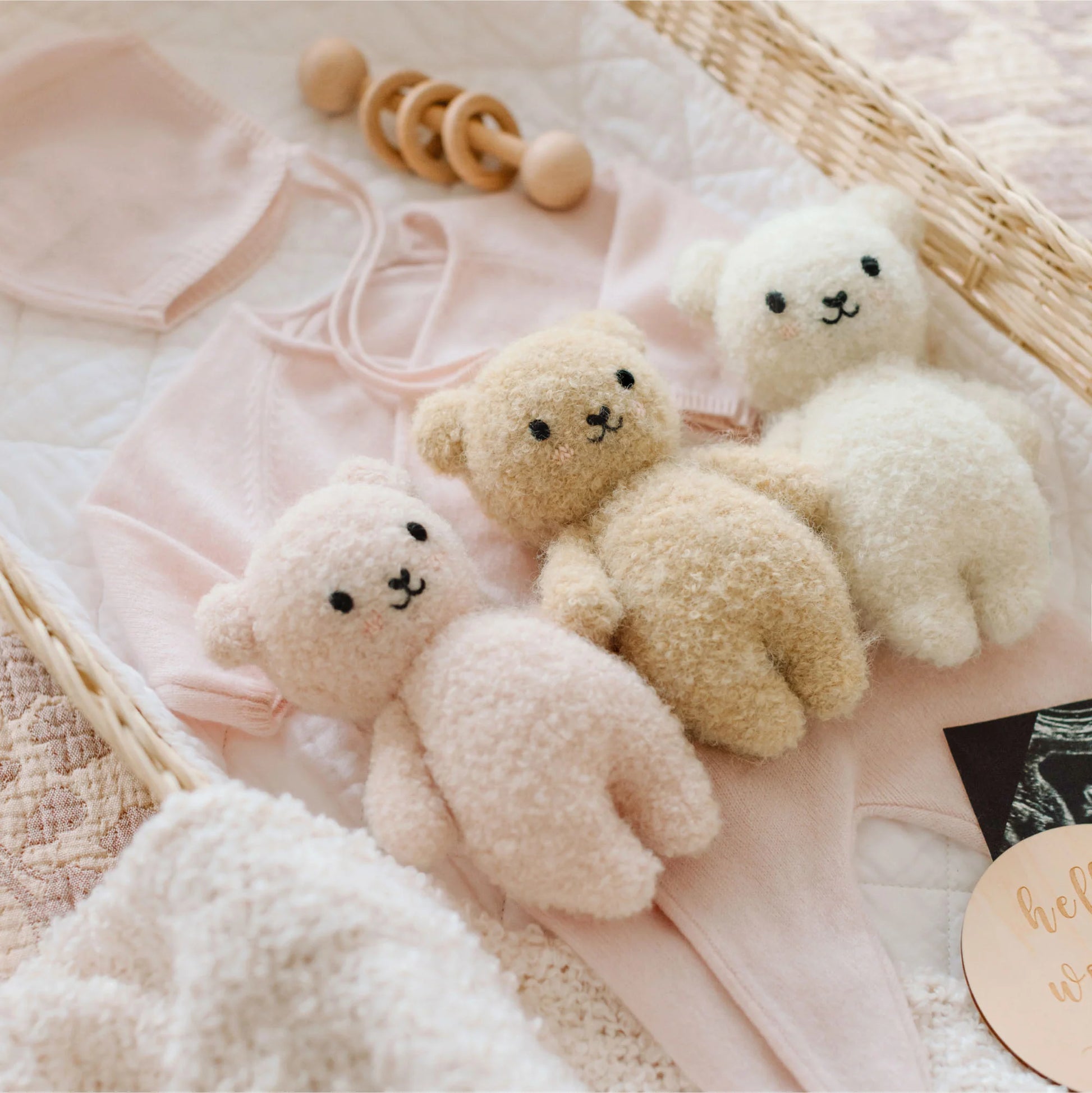 The Cuddle + Kind Baby Boucle Bear - Strawberry, lovingly crafted from soft Peruvian cotton yarn, is featured in a charming gift basket alongside baby clothes, a wooden rattle, and a greeting card.