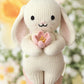 A hand holds the Baby Bunny - Blooming Tulip by Cuddle + Kind—an heirloom-quality, cream-colored bunny plush made from handcrafted Peruvian cotton, clutching a pink tulip, with a blurred floral background behind.