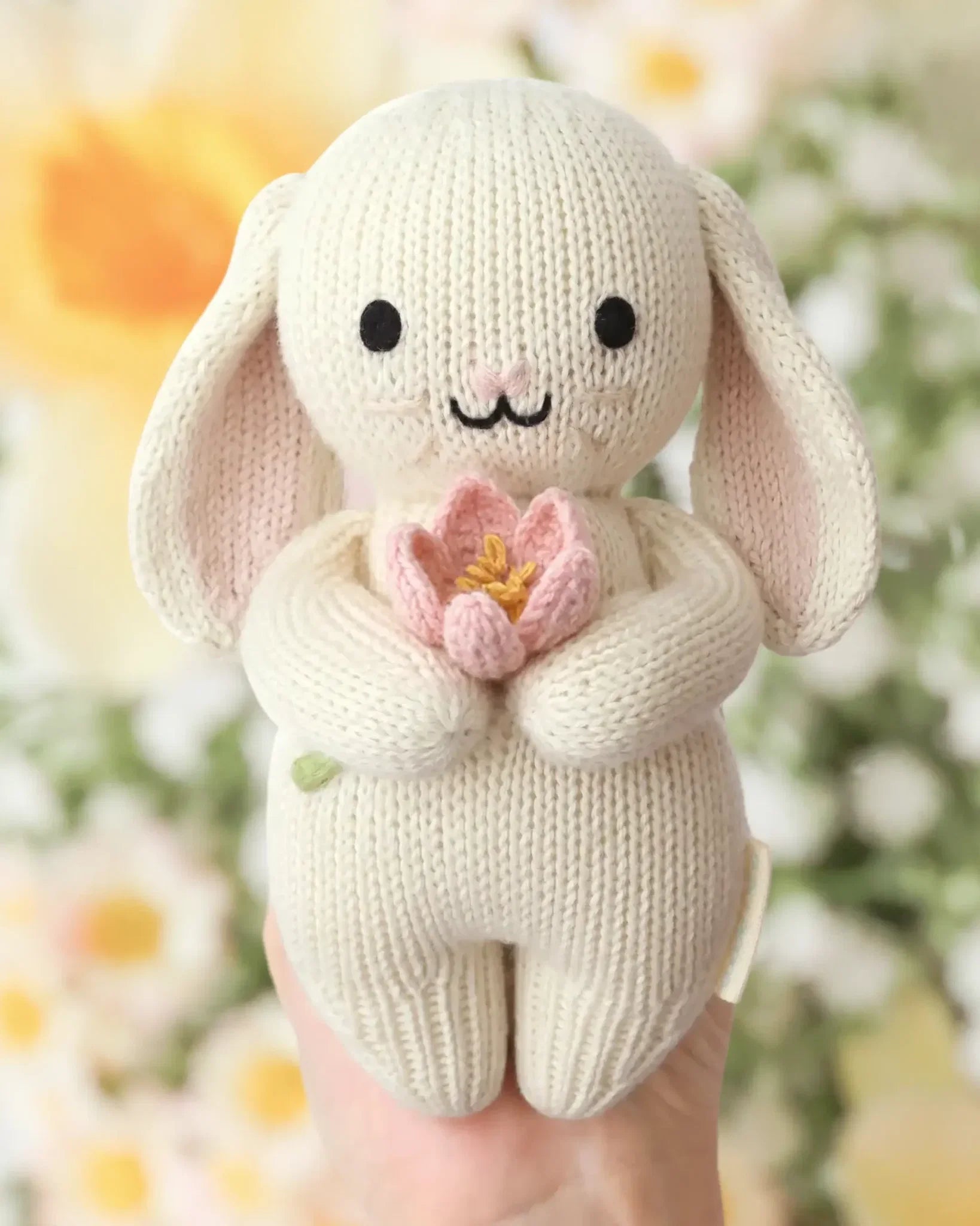 A hand holds the Baby Bunny - Blooming Tulip by Cuddle + Kind—an heirloom-quality, cream-colored bunny plush made from handcrafted Peruvian cotton, clutching a pink tulip, with a blurred floral background behind.
