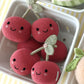 Five “Baby Cherries” knitted plush toys from Cuddle + Kind, each with a smiling face and green stem and handcrafted from cotton yarn, are arranged in a white tray on a striped yellow and white surface.