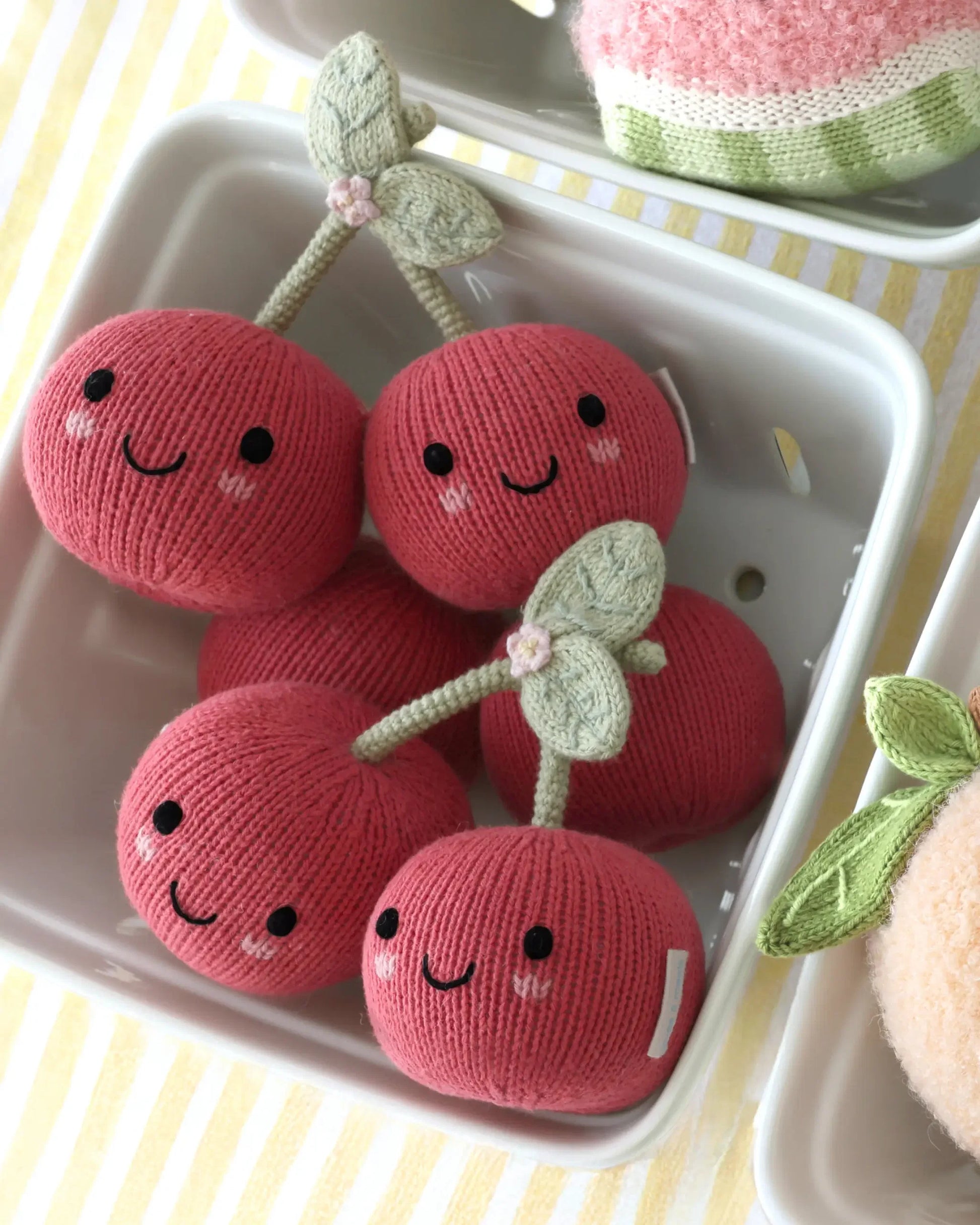 Five “Baby Cherries” knitted plush toys from Cuddle + Kind, each with a smiling face and green stem and handcrafted from cotton yarn, are arranged in a white tray on a striped yellow and white surface.