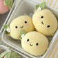 Three Cuddle + Kind Baby Lemon plush toys—yellow, knitted, and handcrafted from Peruvian cotton with embroidered smiling faces and green leaf accents—sit in a white dish on a striped tablecloth.