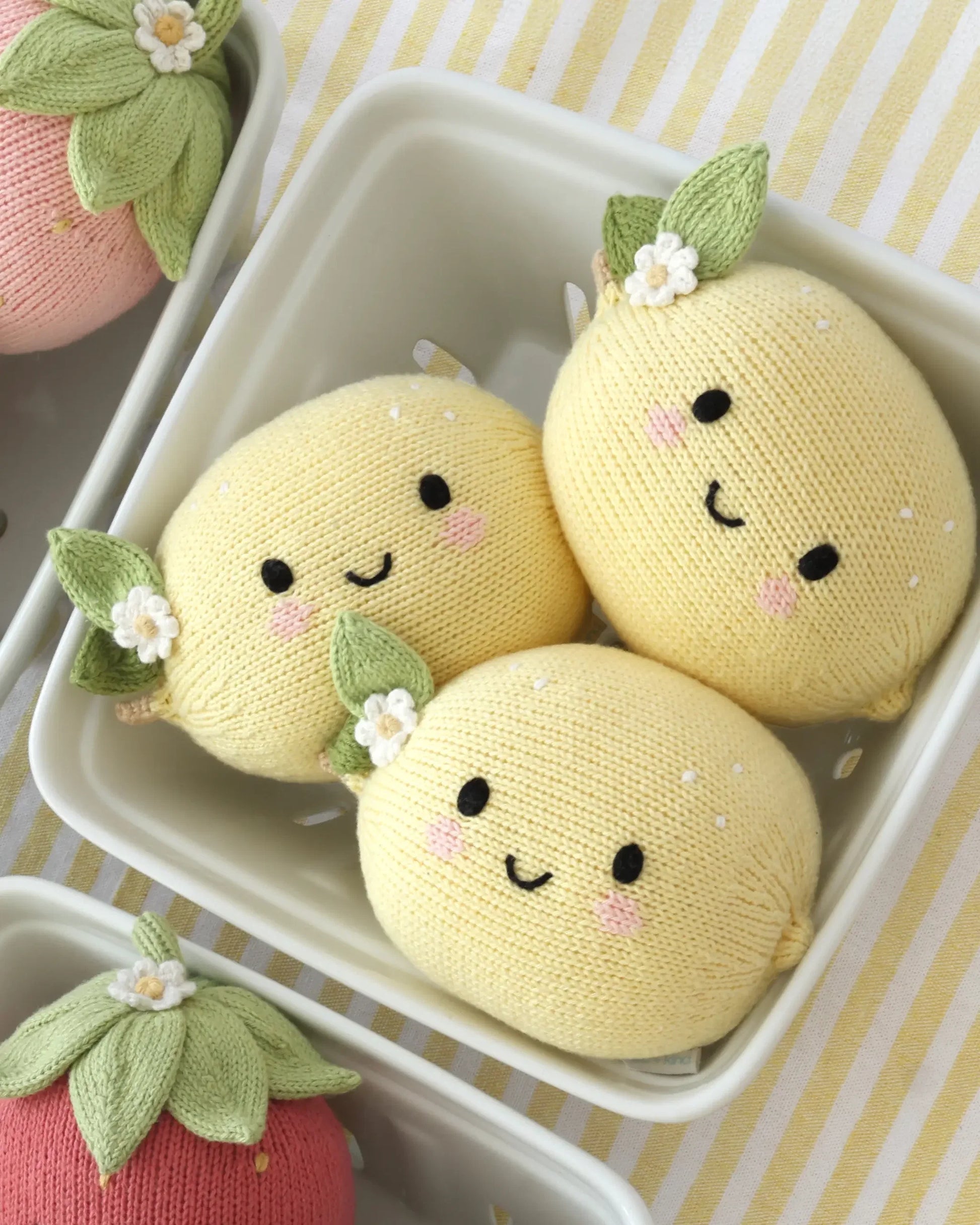Three Cuddle + Kind Baby Lemon plush toys—yellow, knitted, and handcrafted from Peruvian cotton with embroidered smiling faces and green leaf accents—sit in a white dish on a striped tablecloth.