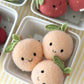 Three Cuddle + Kind “Baby Peach” plush toys—with smiling faces and green leaves, each handcrafted from soft Peruvian cotton yarn—sit in a small white basket on a striped yellow and white tablecloth.
