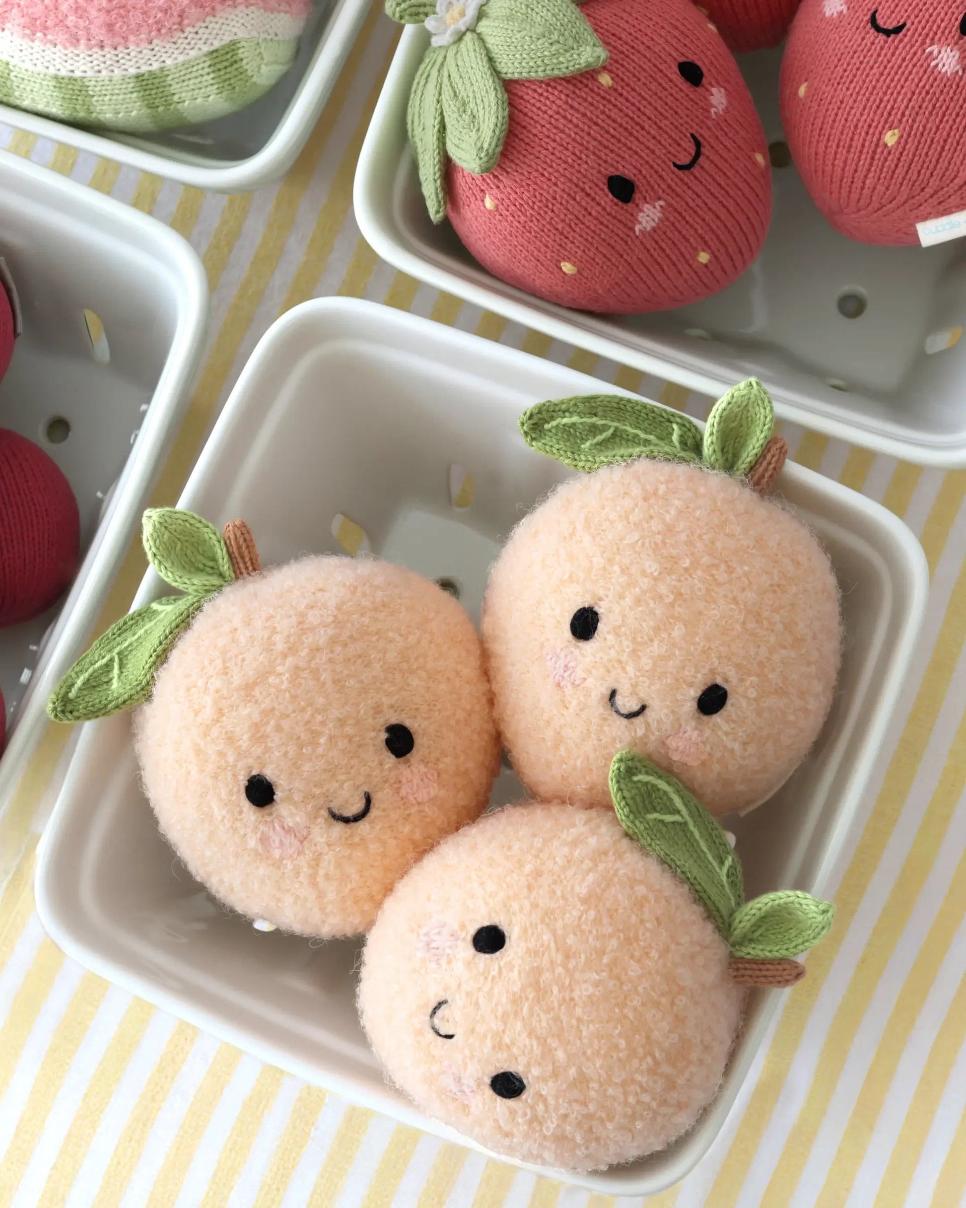 Three Cuddle + Kind “Baby Peach” plush toys—with smiling faces and green leaves, each handcrafted from soft Peruvian cotton yarn—sit in a small white basket on a striped yellow and white tablecloth.