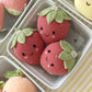 Three Baby Strawberry plushies from Cuddle + Kind, handcrafted with care from Peruvian cotton yarn and embellished with embroidered faces and green fabric leaves, sit in a white dish surrounded by other plush fruits—including an heirloom-quality baby pumpkin—on a yellow-and-white striped surface.