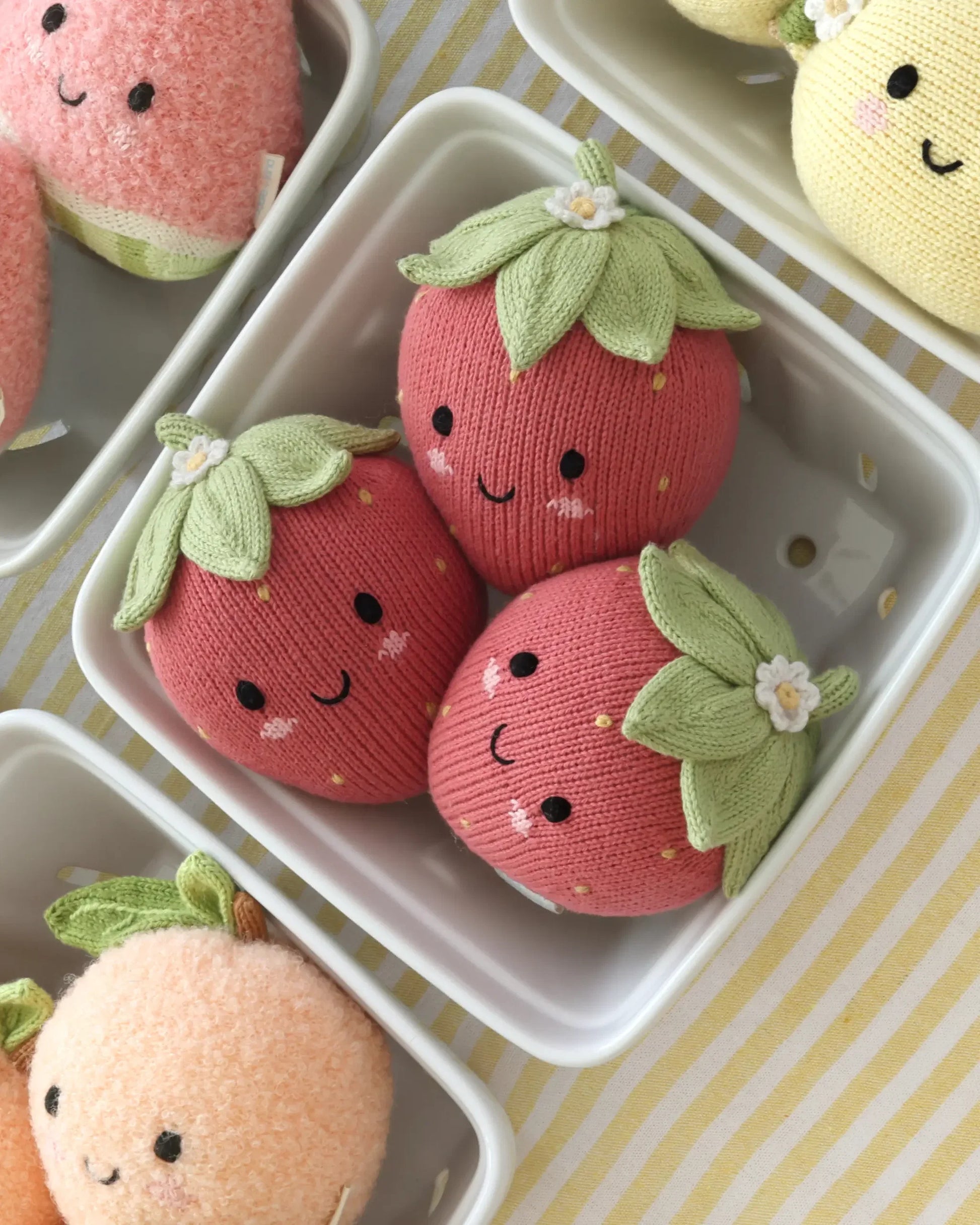 Three Baby Strawberry plushies from Cuddle + Kind, handcrafted with care from Peruvian cotton yarn and embellished with embroidered faces and green fabric leaves, sit in a white dish surrounded by other plush fruits—including an heirloom-quality baby pumpkin—on a yellow-and-white striped surface.