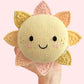 A hand holds the Cuddle + Kind “Baby Sun”—a handcrafted cotton toy with a smiling face and cheerful yellow and pink rays—set against a pale pink background, making it an adorable companion sure to brighten any nursery.