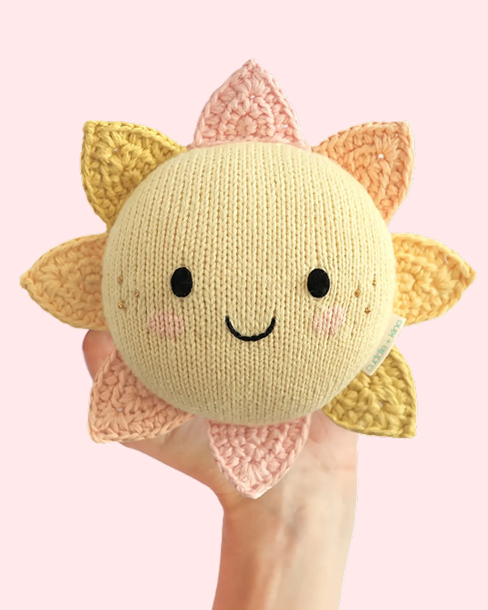 A hand holds the Cuddle + Kind “Baby Sun”—a handcrafted cotton toy with a smiling face and cheerful yellow and pink rays—set against a pale pink background, making it an adorable companion sure to brighten any nursery.