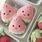Three plush Baby Watermelon slices by Cuddle + Kind, each featuring a cheerful smile, are displayed in a white tray surrounded by other charming plush fruit toys—including an adorable heirloom baby pumpkin—on a striped yellow and white surface. Each soft baby toy is lovingly handcrafted from the finest Peruvian cotton, ensuring both comfort and charm for little ones.
