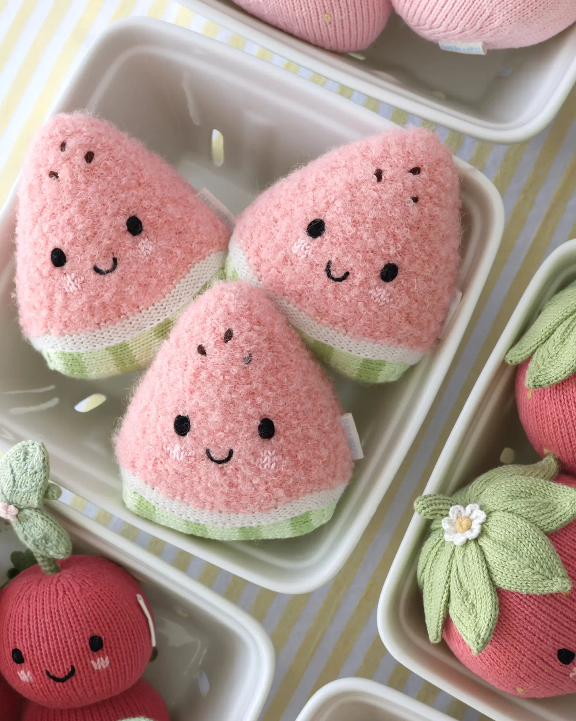 Three plush Baby Watermelon slices by Cuddle + Kind, each featuring a cheerful smile, are displayed in a white tray surrounded by other charming plush fruit toys—including an adorable heirloom baby pumpkin—on a striped yellow and white surface. Each soft baby toy is lovingly handcrafted from the finest Peruvian cotton, ensuring both comfort and charm for little ones.