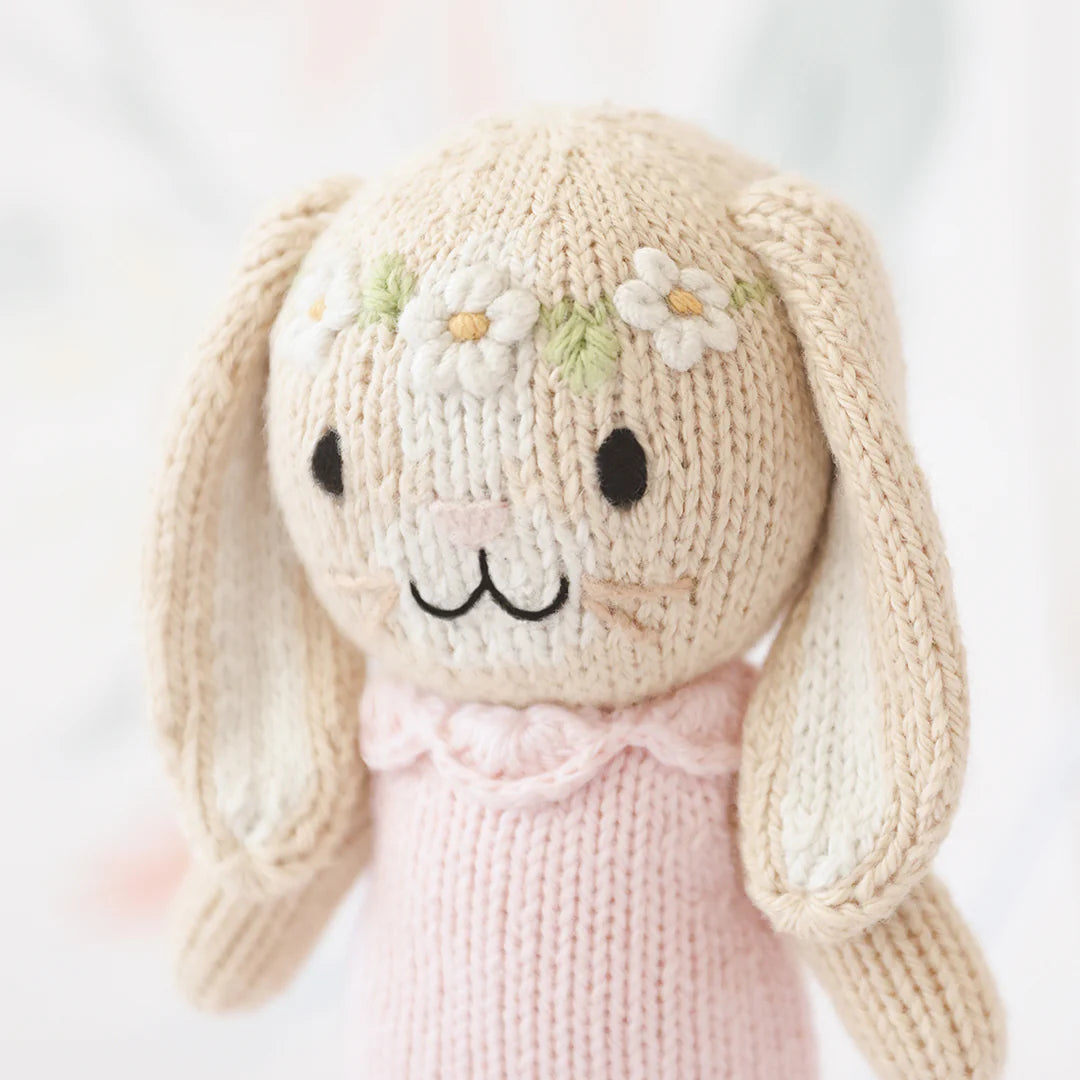 Tiny Hannah the Bunny Blush by Cuddle + Kind is an artisan-made, fair trade hand-knit bunny doll with long floppy ears, black eyes, floral accents on its head, and a pale pink sweater.