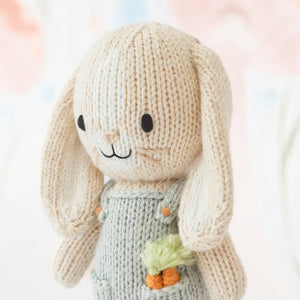 Tiny Henry the Bunny by Cuddle + Kind is a hand-knit doll in light gray overalls with an embroidered carrot, posed on a soft pastel background. Part of the cuddle+kind tiny collection.