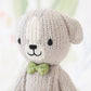 Close-up of a hand-knit Tiny Noah the Dog doll by Cuddle + Kind, featuring a light gray body, black stitched facial features, and a green bow tie from the Tiny Collection.