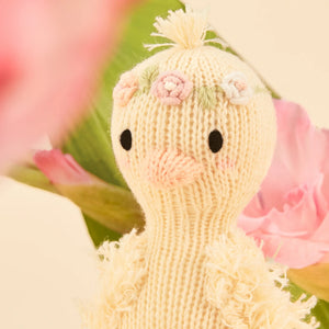 Tiny Flora the Duckling Mauve by Cuddle + Kind is a hand-knit plush bird with embroidered flowers on its head, black eyes, and a pink beak, shown in front of pink flowers and green leaves.