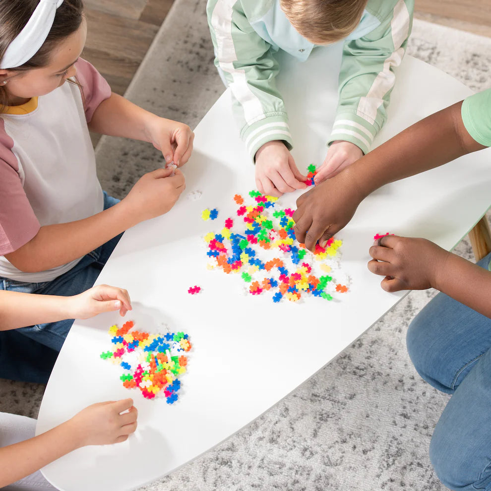 Four children sit around a white table on a grey carpet, assembling Plus Plus Tube - 240 Piece Neon Mix—colorful STEM toys that inspire creative play and help develop fine motor skills.
