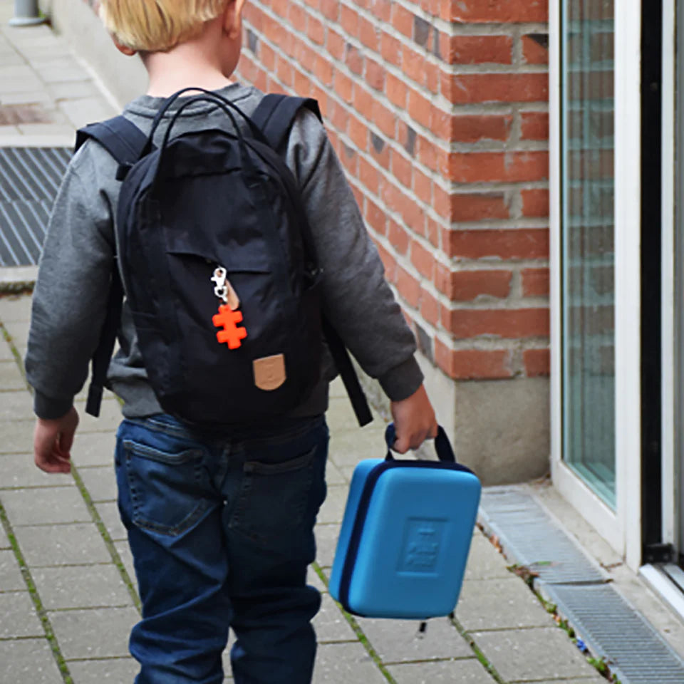 A young child with a black backpack and red puzzle piece keychain walks on a sidewalk by a brick building, carrying the Plus Plus Travel Case for creative play.