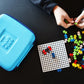 Someone assembles colorful Plus-Plus pieces on a white grid beside a closed blue Plus Plus Travel Case, encouraging hands-on STEM learning and creative fun.