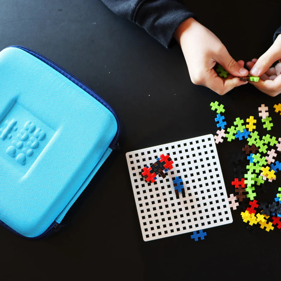 Someone assembles colorful Plus-Plus pieces on a white grid beside a closed blue Plus Plus Travel Case, encouraging hands-on STEM learning and creative fun.