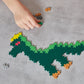 A hand places a green piece onto a partially built pixelated dinosaur puzzle from the Plus Plus Learn To Build - Dinosaurs set, on a gray surface.
