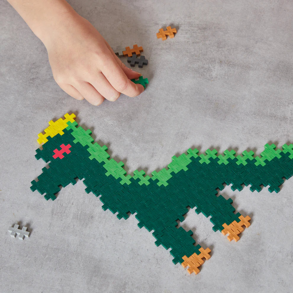 A hand places a green piece onto a partially built pixelated dinosaur puzzle from the Plus Plus Learn To Build - Dinosaurs set, on a gray surface.
