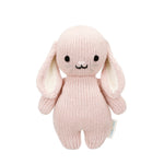 The Cuddle + Kind Rose Bunny - Baby 7" is a hand-knit stuffed bunny, crafted from soft Peruvian cotton yarn. Featuring a gentle pink color, floppy ears, black eyes, and a small smiling mouth, this heirloom-quality plush stands upright against a white background—perfect for any baby animal collection.