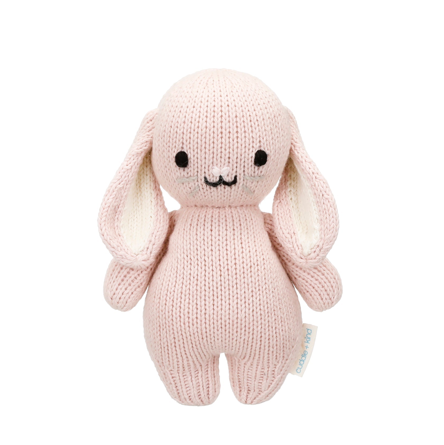 The Cuddle + Kind Rose Bunny - Baby 7" is a hand-knit stuffed bunny, crafted from soft Peruvian cotton yarn. Featuring a gentle pink color, floppy ears, black eyes, and a small smiling mouth, this heirloom-quality plush stands upright against a white background—perfect for any baby animal collection.