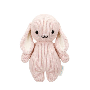 The Cuddle + Kind Rose Bunny - Baby 7" is a hand-knit stuffed bunny, crafted from soft Peruvian cotton yarn. Featuring a gentle pink color, floppy ears, black eyes, and a small smiling mouth, this heirloom-quality plush stands upright against a white background—perfect for any baby animal collection.