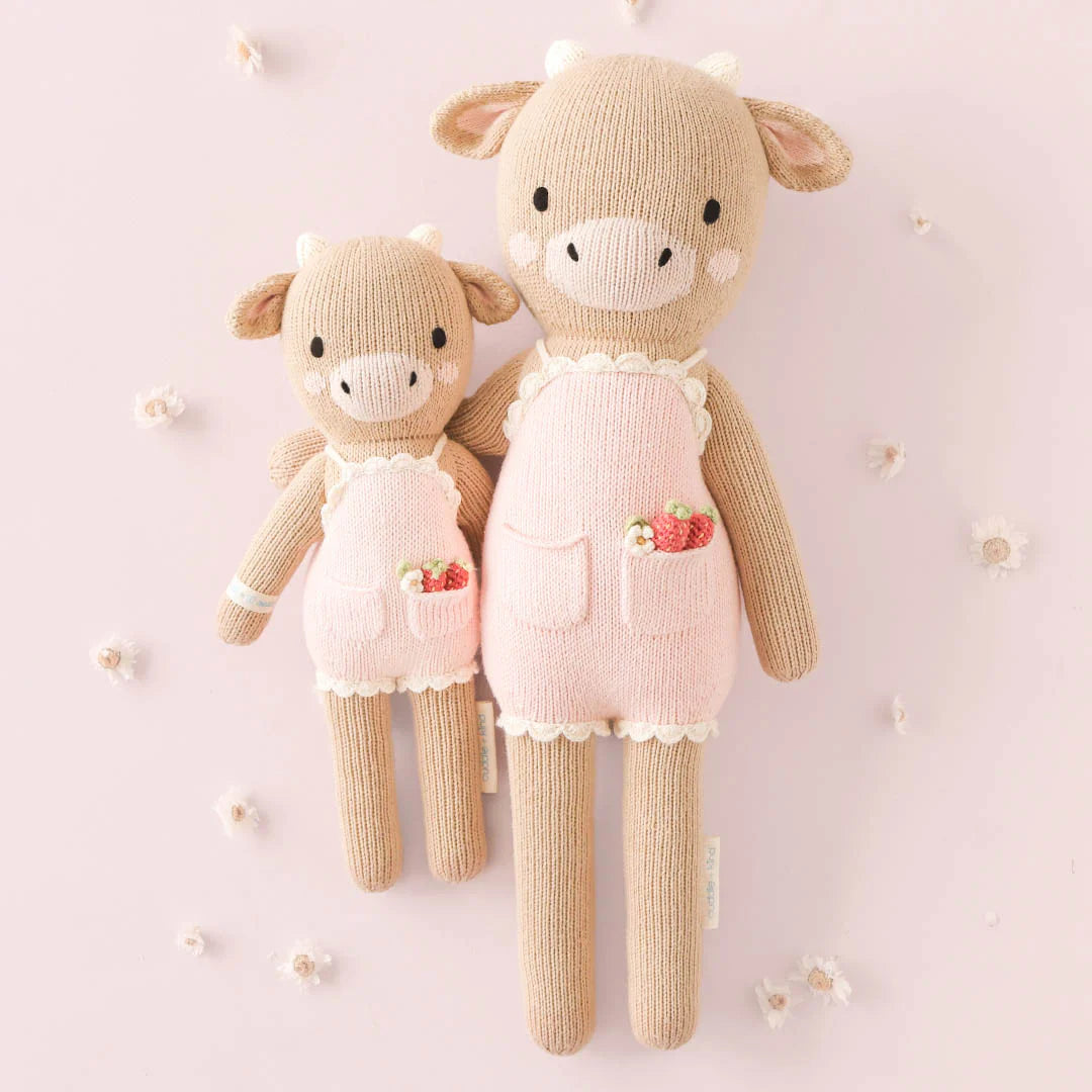 Ava the Cow Powder Pink (Regular 20") by Cuddle + Kind stands on a light pink background with white flowers, looking ready to celebrate—perfect for adding a cheerful touch to any birthday or special occasion.