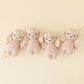 Four “Baby Bunny - Rose w/ Ivory Floral” plush toys by Cuddle + Kind are arranged in a row on a beige background, each handcrafted from soft baby toy cotton yarn and adorned with delicate ivory flower crowns, with small white flowers scattered nearby.