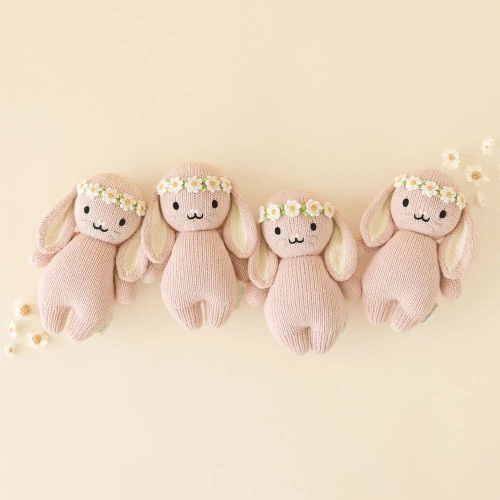 Four “Baby Bunny - Rose w/ Ivory Floral” plush toys by Cuddle + Kind are arranged in a row on a beige background, each handcrafted from soft baby toy cotton yarn and adorned with delicate ivory flower crowns, with small white flowers scattered nearby.