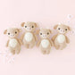 Four identical Cuddle + Kind Baby Cow toys—hand-knitted in beige and white from handcrafted cotton yarn—are arranged in a row against a light pink background with scattered white flowers, creating a charming soft toy set perfect for little hands.