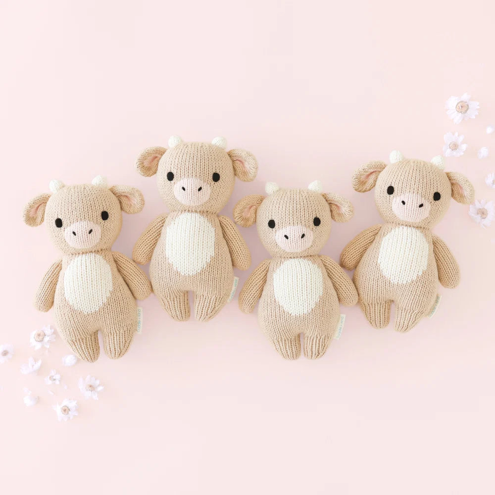 Four identical Cuddle + Kind Baby Cow toys—hand-knitted in beige and white from handcrafted cotton yarn—are arranged in a row against a light pink background with scattered white flowers, creating a charming soft toy set perfect for little hands.