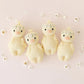 Four Cuddle + Kind Baby Duckling Ivory Floral dolls with flower crowns lie side by side on a pale surface, surrounded by small white flowers.