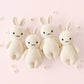 Four cream-colored Baby Rabbit - Oatmeal dolls by Cuddle + Kind, each hand-knit with embroidered faces, are arranged side by side on a light pink background with small floral decorations.